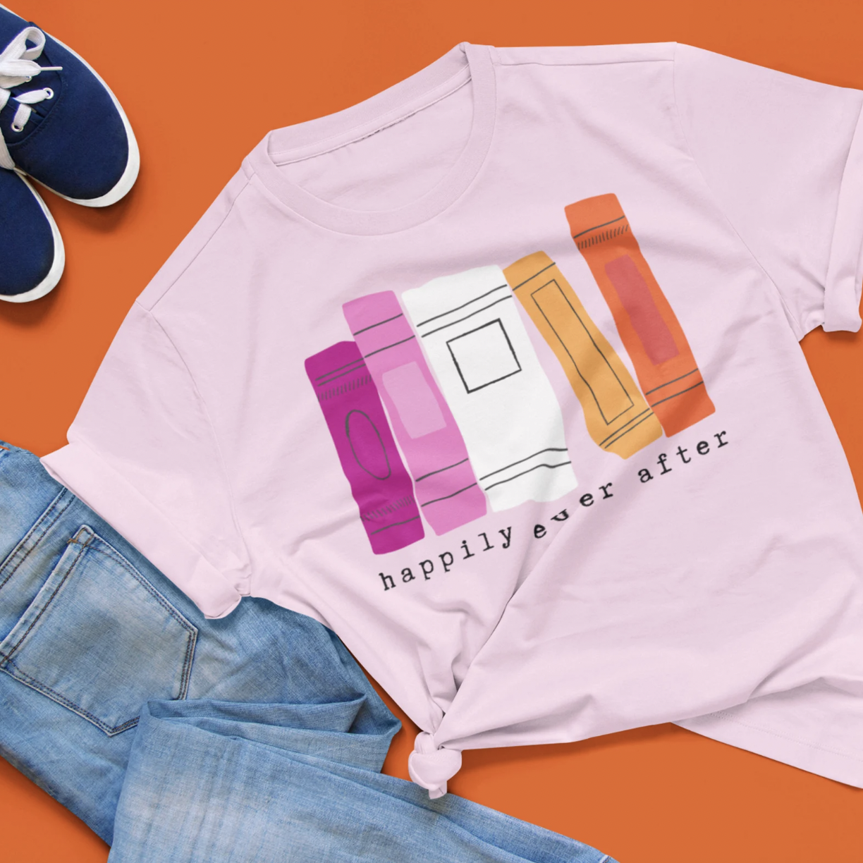 Image of a light pink shirt with an illustration of book spines in the lesbian pride flag colors with the text "happily ever after"