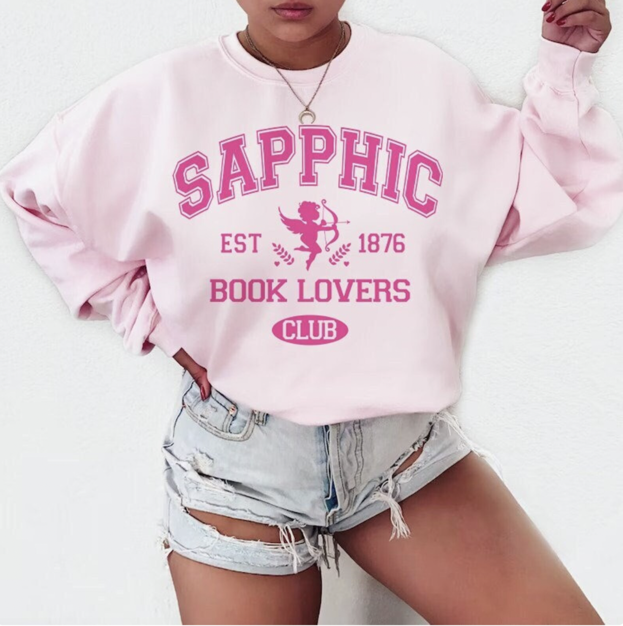 Image of someone in a light pink sweatshirt with hot pink text that says "Sapphic Book Lovers Club Est. 1876"