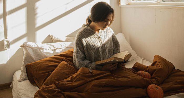 fair-skinned woman reading a book in bed with a brown comforter and pumpkins near her